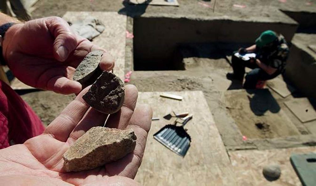 Close up photo of a worker's hands holding excavated materials. Another worker is working