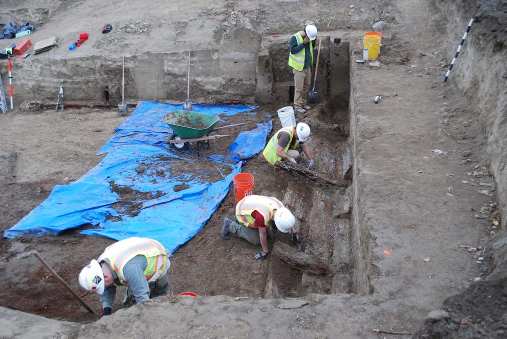 Photo of workers digging in a site. There is a large blue tarp where workers are digging