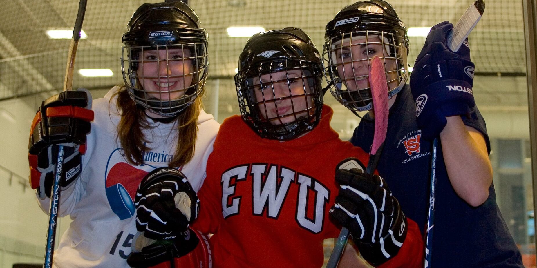 Baitlyn, Kayla, and Daitlin pose, wearing hockey gear, in front of the rec center's ice rink