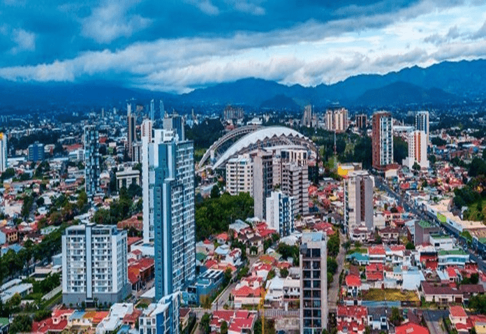 Digital image San Jose Costa Rica, several tall sky scrapers and a stadium, on a blue day with a white streak of clouds