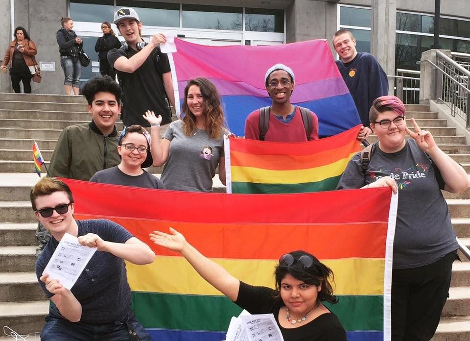 Students smiling on the JFK Library steps holding pride and bisexuality flags