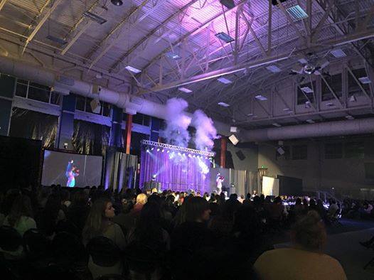 Drag show event at EWU. Stage lit without purple lights with projects on either side. There are many people in front in the crowd