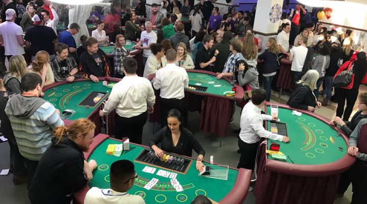 People at four casino card tables playing various games. There are four dealers giviing out cards