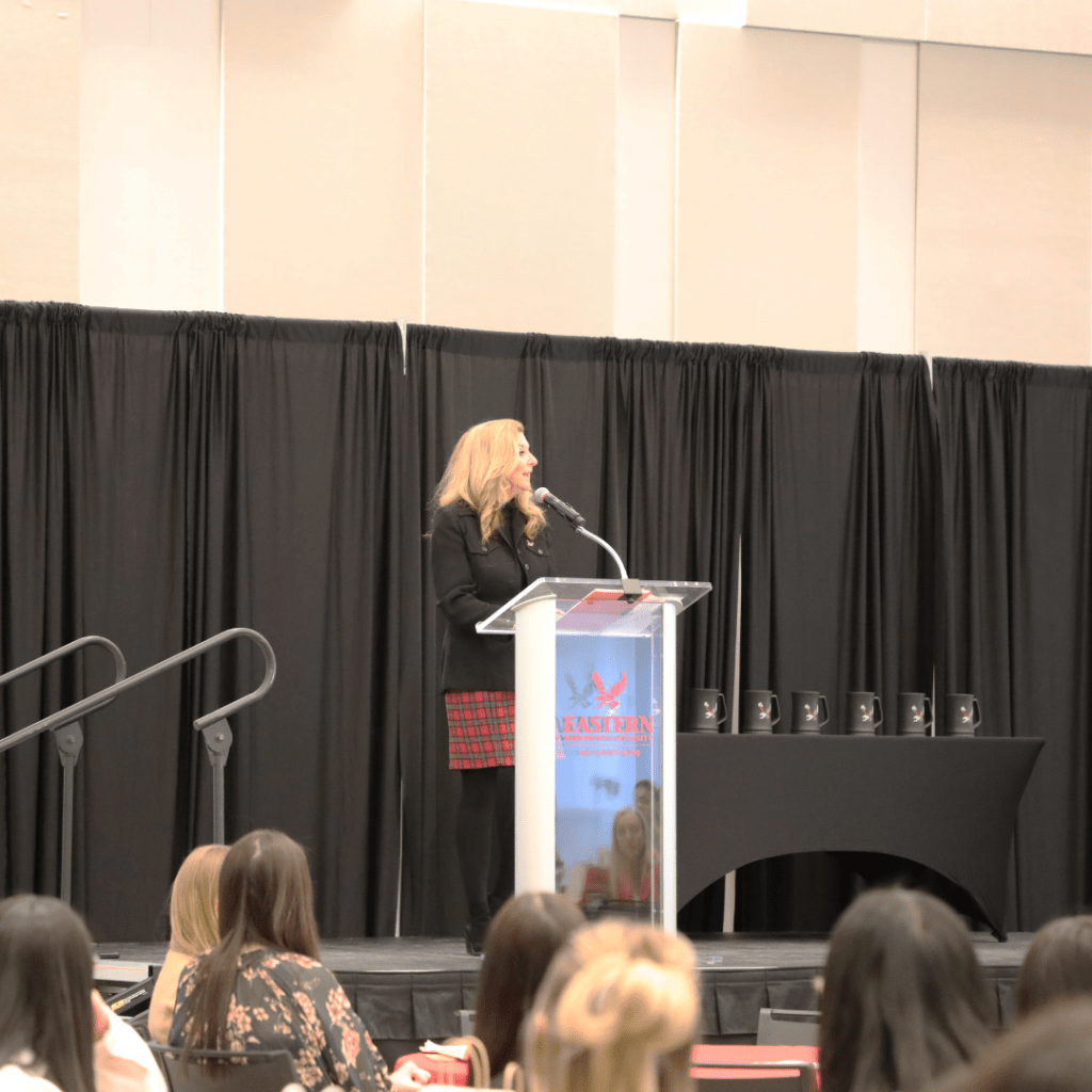 Women speaking at the SFL professional summit in front of a crowd on a stage. This is in the Nysether Community Room.