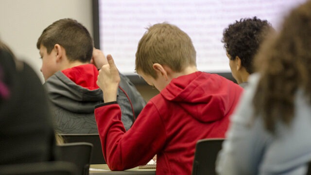 back of student holding their hand up. They are sitting at a desk in a classroom with students